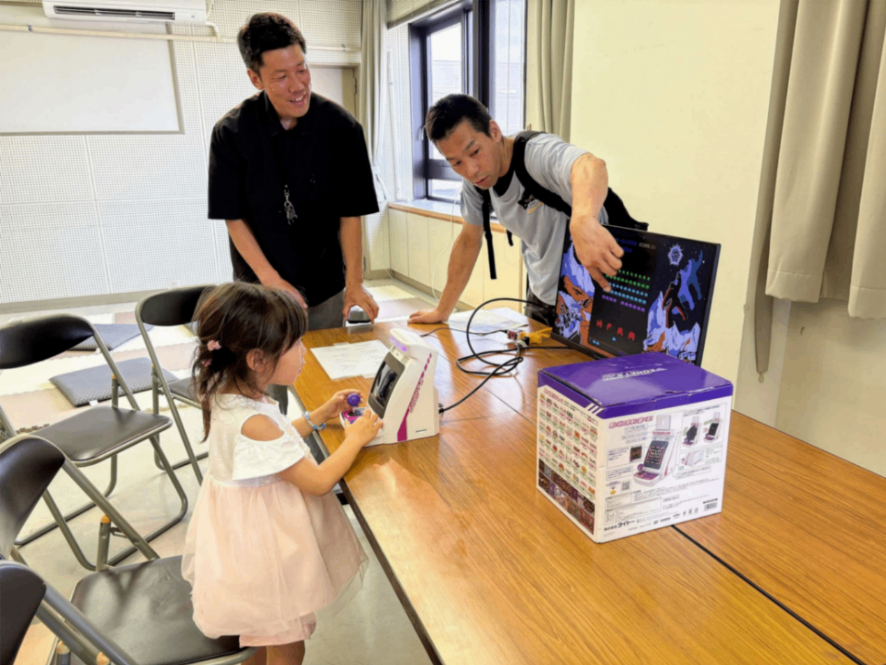 写真：レトロゲームに挑戦する小さな女の子。画面を指差してアドバイスをする男性や、その様子を笑顔で見守っている男性がいます。