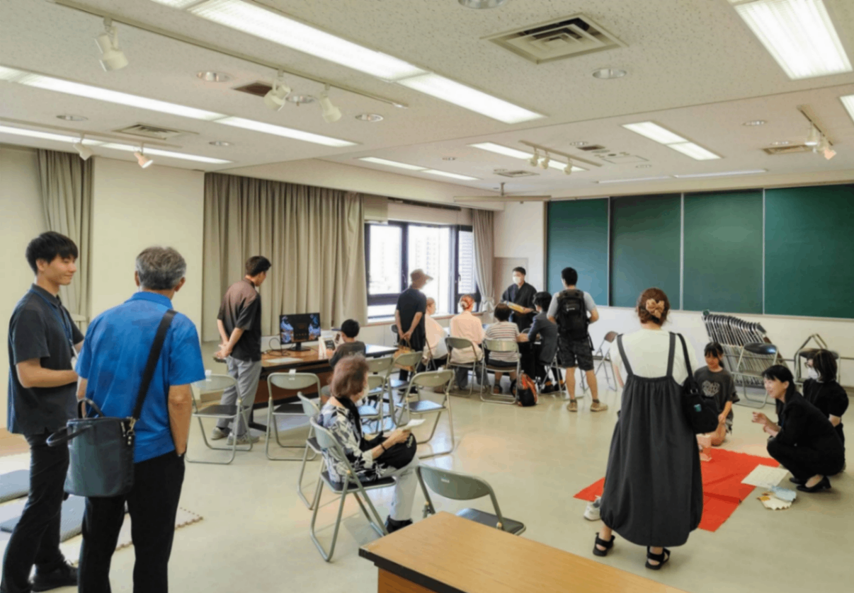 明るい会議室で、数人の参加者が椅子に座ったり立ったりして交流している様子。部屋の奥には黒板があり、手前には数人が集まって談笑している。