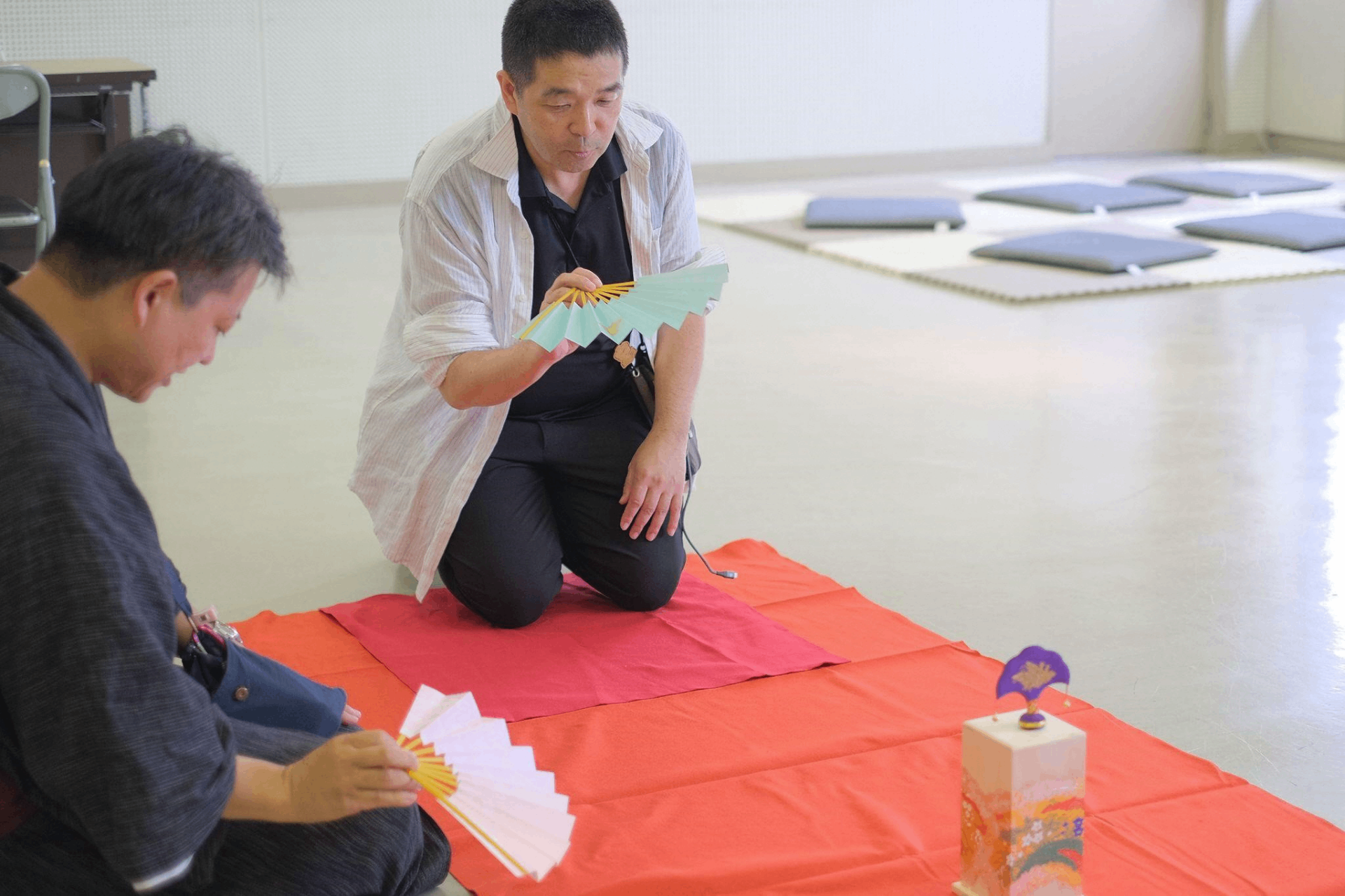 室内で正座をした二人の男性が、それぞれ扇子を手に持ち、赤い布が敷かれた床の上の的を見つめている。