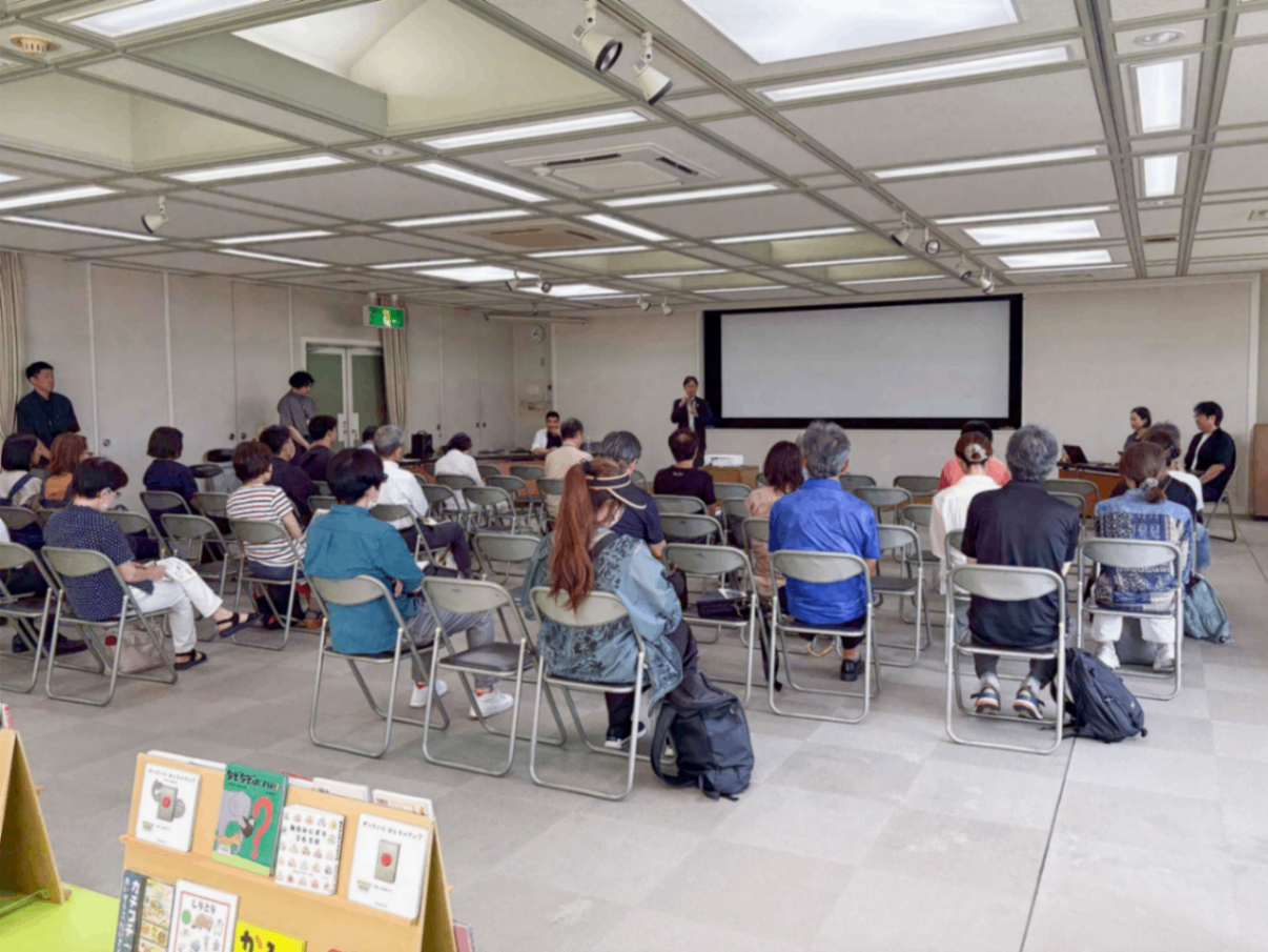 写真：会場の様子。前方にスクリーンがあり、スーツ姿の男性がマイクを持って話しています。参加者はパイプ椅子に座って聞いています。後方には絵本のコーナーがあります。
